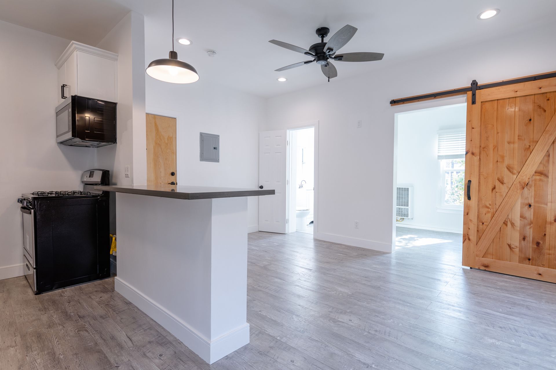 Modern, bright studio apartment with kitchen area, bar, barn door, and ceiling fan. White and wood tones.