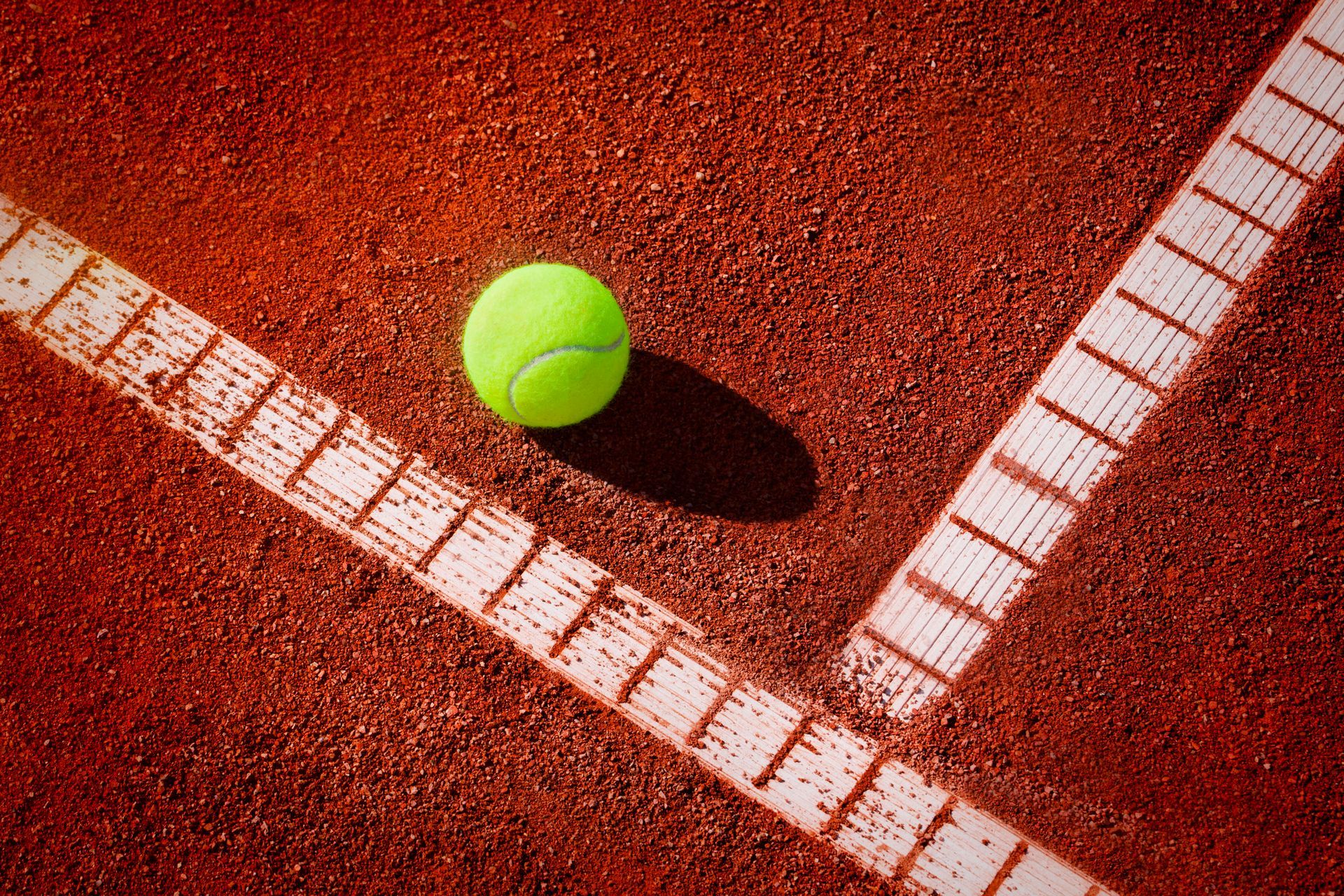 Una pallina da tennis giallo neon appoggiata su un campo da tennis in terra battuta rossa, vicino a una linea bianca dipinta.