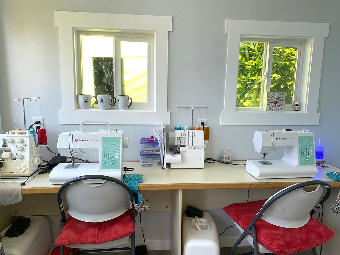 Two sewing machines are sitting on a desk in front of two windows.
