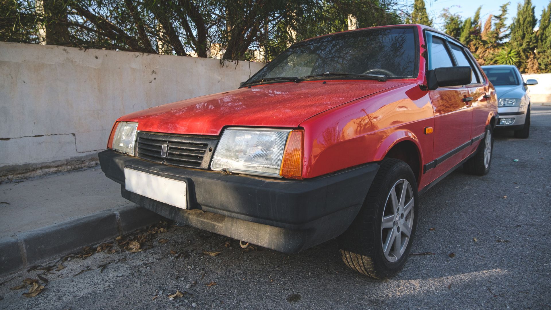 Red hatchback with a black bumper parked by a curb beside a wall, front damage visible