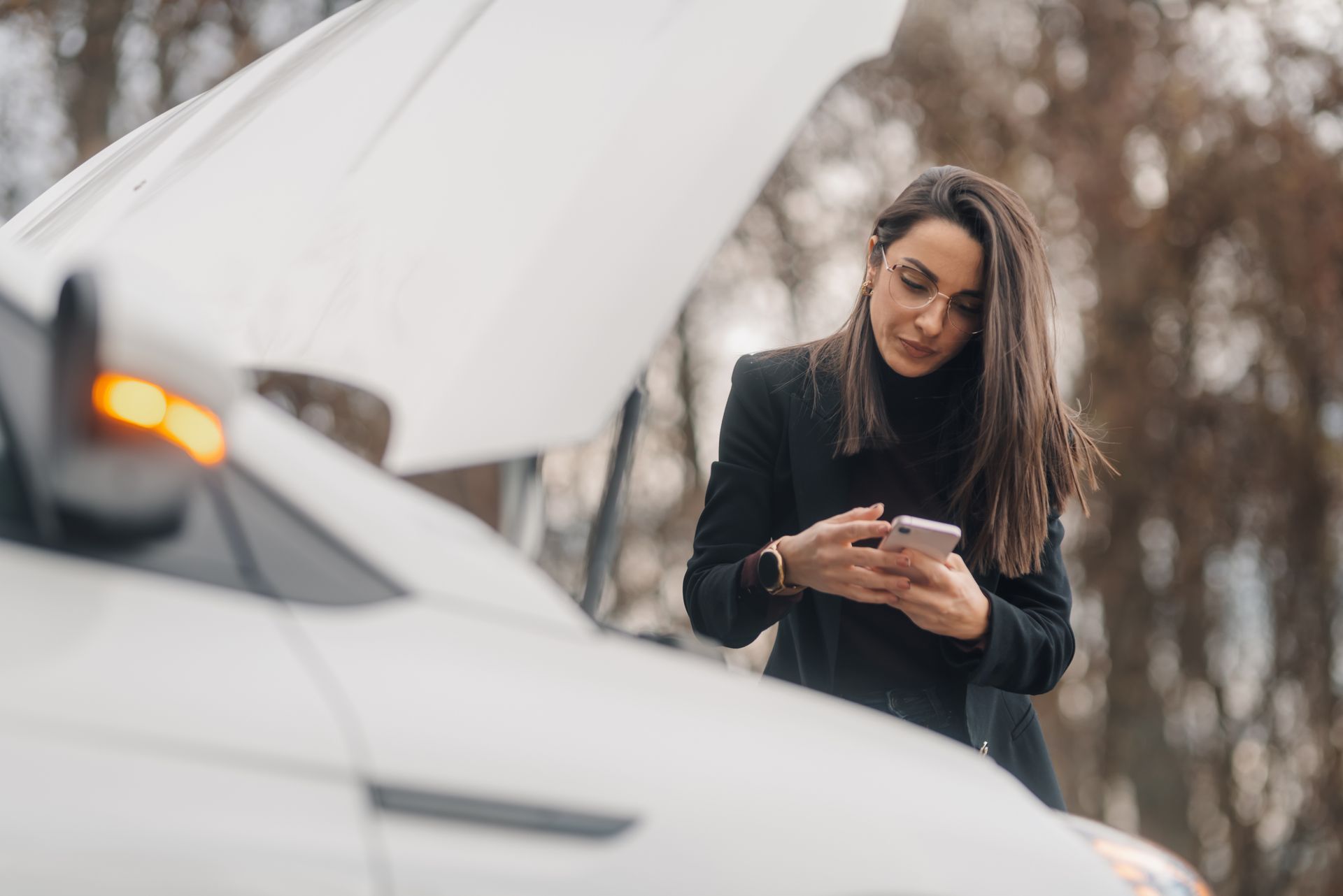 Woman checking her phone beside a white car with the hood open on a roadside