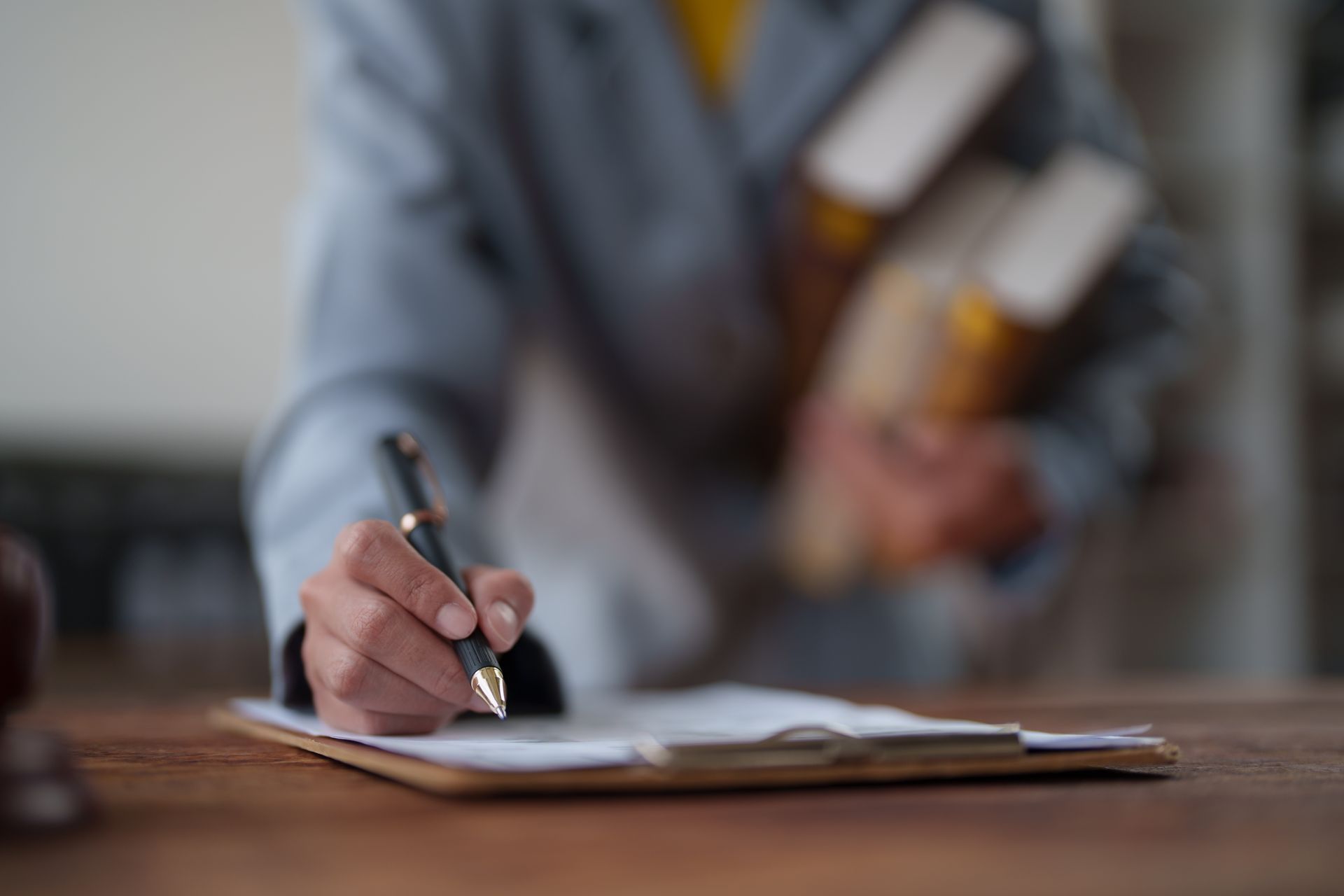 Hand writing on a clipboard at a desk, with books blurred in the background