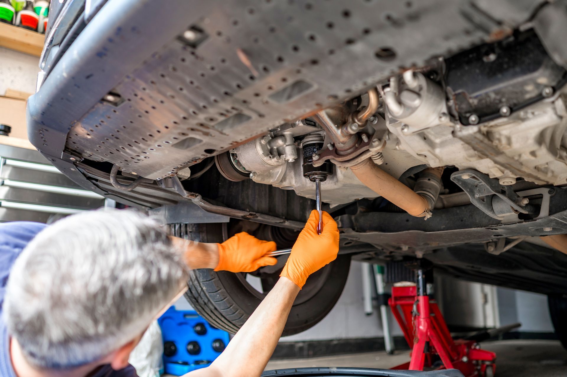 Mechanic under a car inspecting the underside with tools, wearing orange gloves in a garage Mechanic under a car inspecting the underside with tools, wearing orange gloves in a garage
