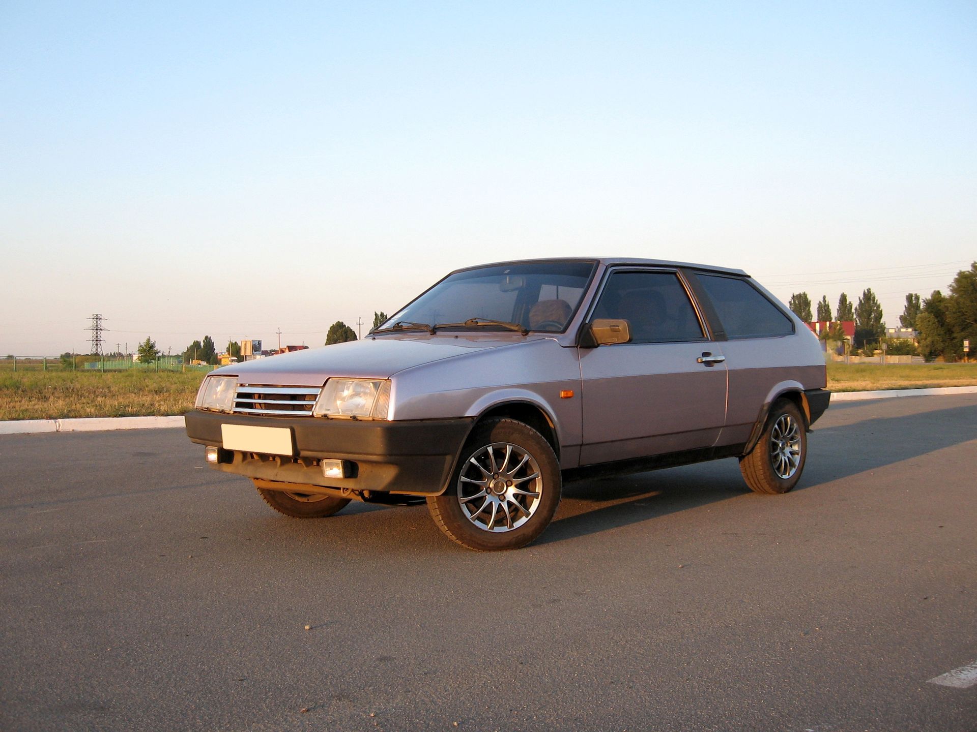 Silver hatchback car parked on a road at sunset with fields and trees in the background Silver hatchback car parked on a road at sunset with fields and trees in the background