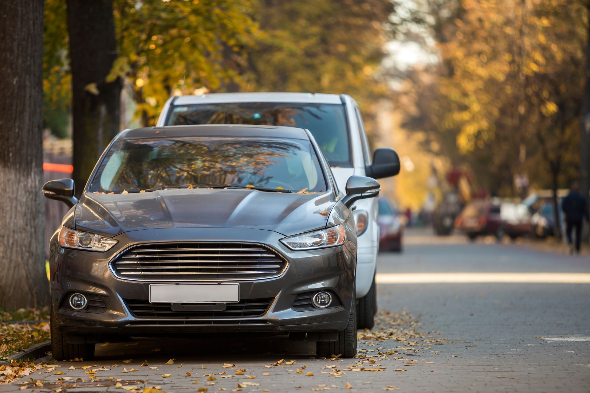 Gray sedan parked on a tree-lined street with autumn leaves, another car behind it.