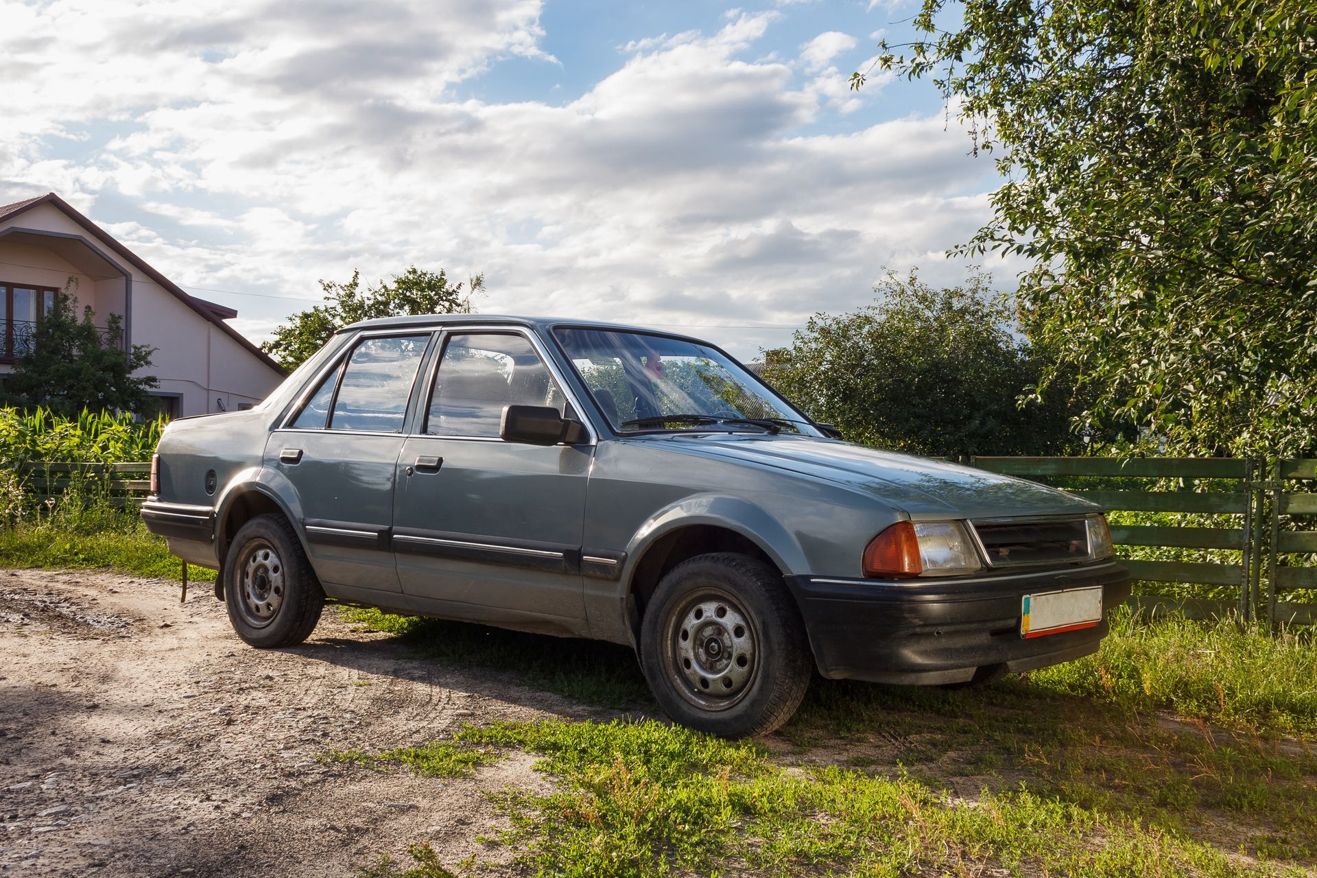 Gray sedan parked on a gravel driveway beside a house, under a cloudy sky