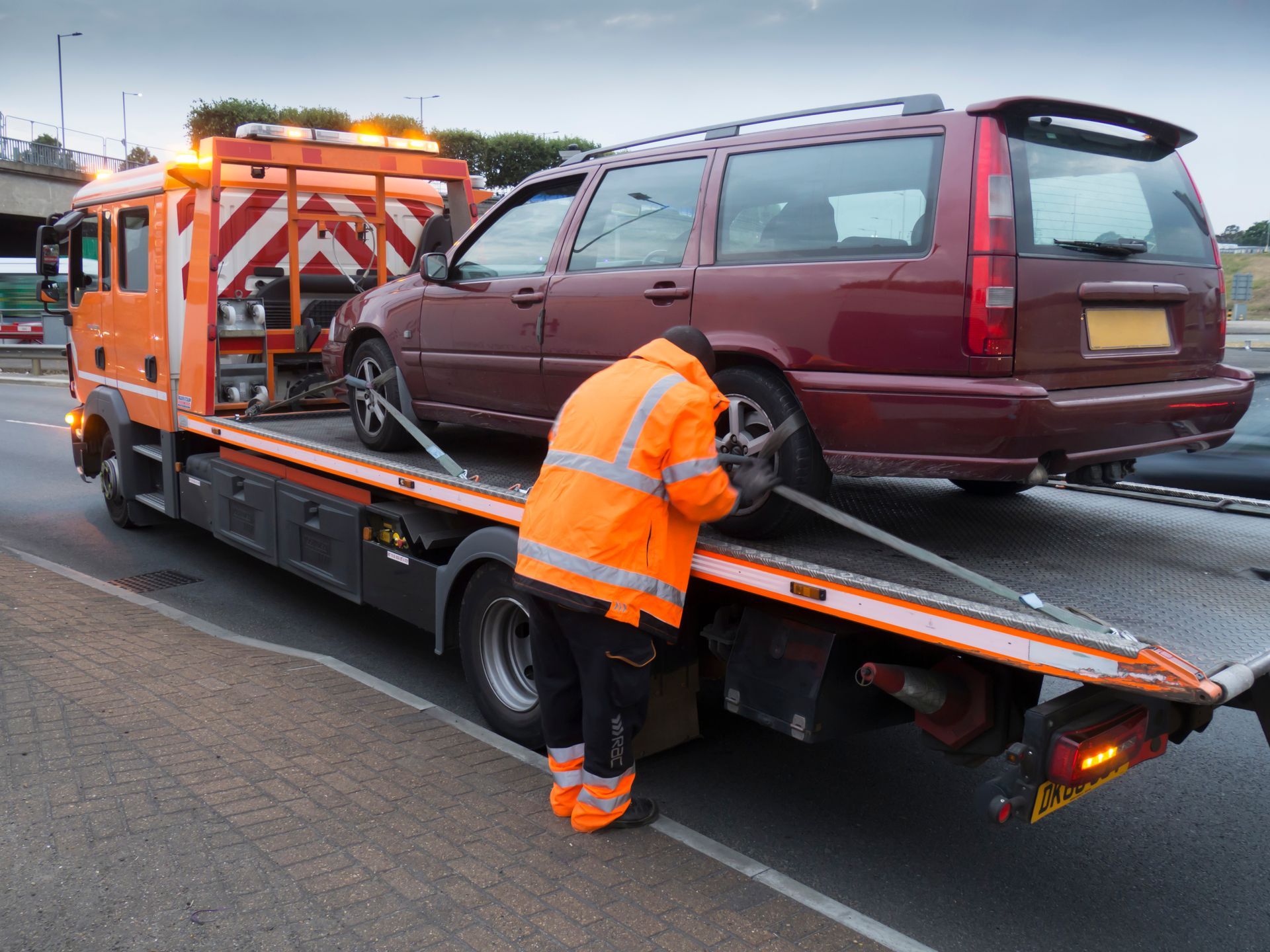 Tow truck operator in orange safety gear securing a maroon SUV on a flatbed at roadside Tow truck operator in orange safety gear securing a maroon SUV on a flatbed at roadside