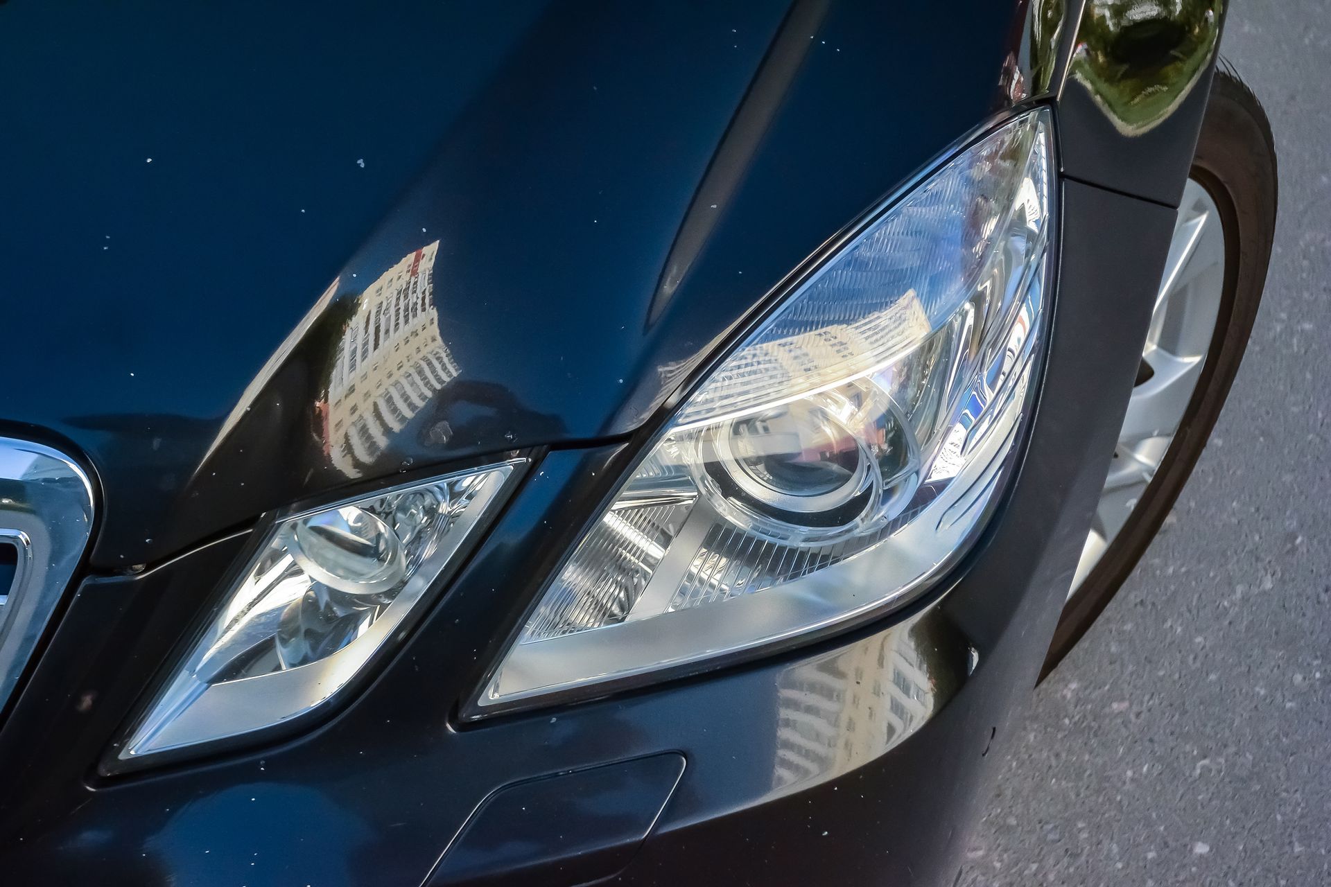 Close-up of a black car’s front headlight and grille reflecting buildings on a sunny street Close-up of a black car’s front headlight and grille reflecting buildings on a sunny street