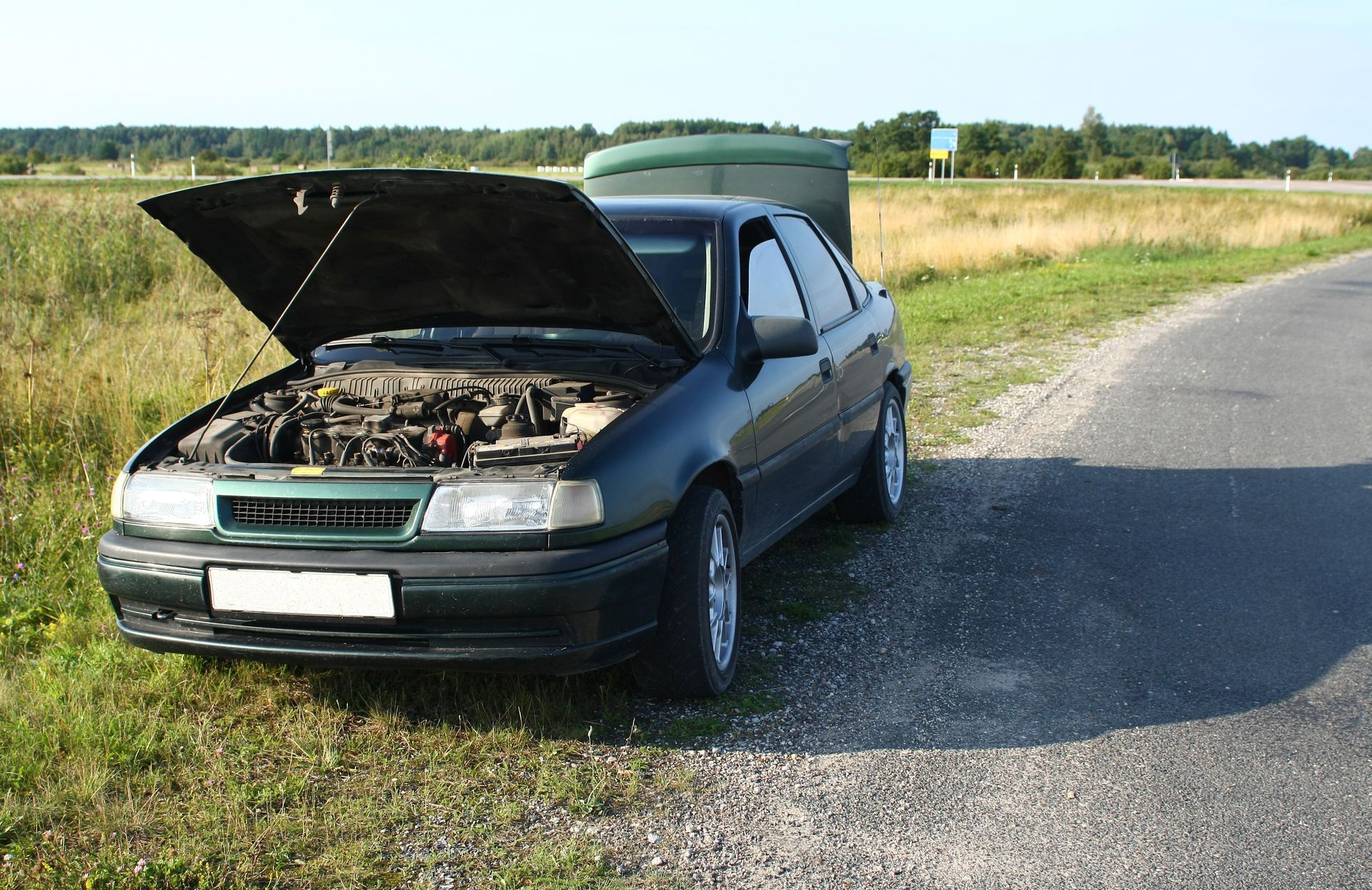 Green car with hood open on a rural roadside, with fields and trees in the background Green car with hood open on a rural roadside, with fields and trees in the background