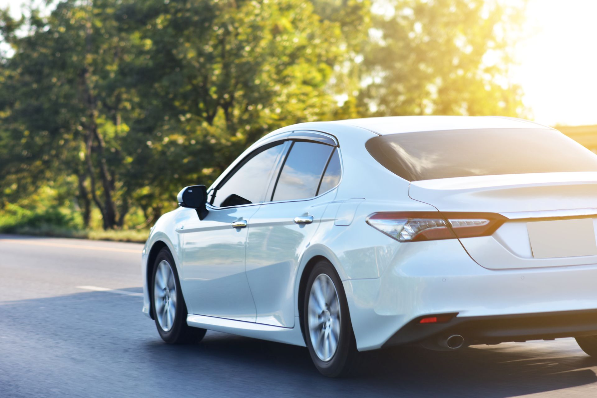 White sedan driving on a sunlit road, viewed from the rear with trees along the roadside White sedan driving on a sunlit road, viewed from the rear with trees along the roadside