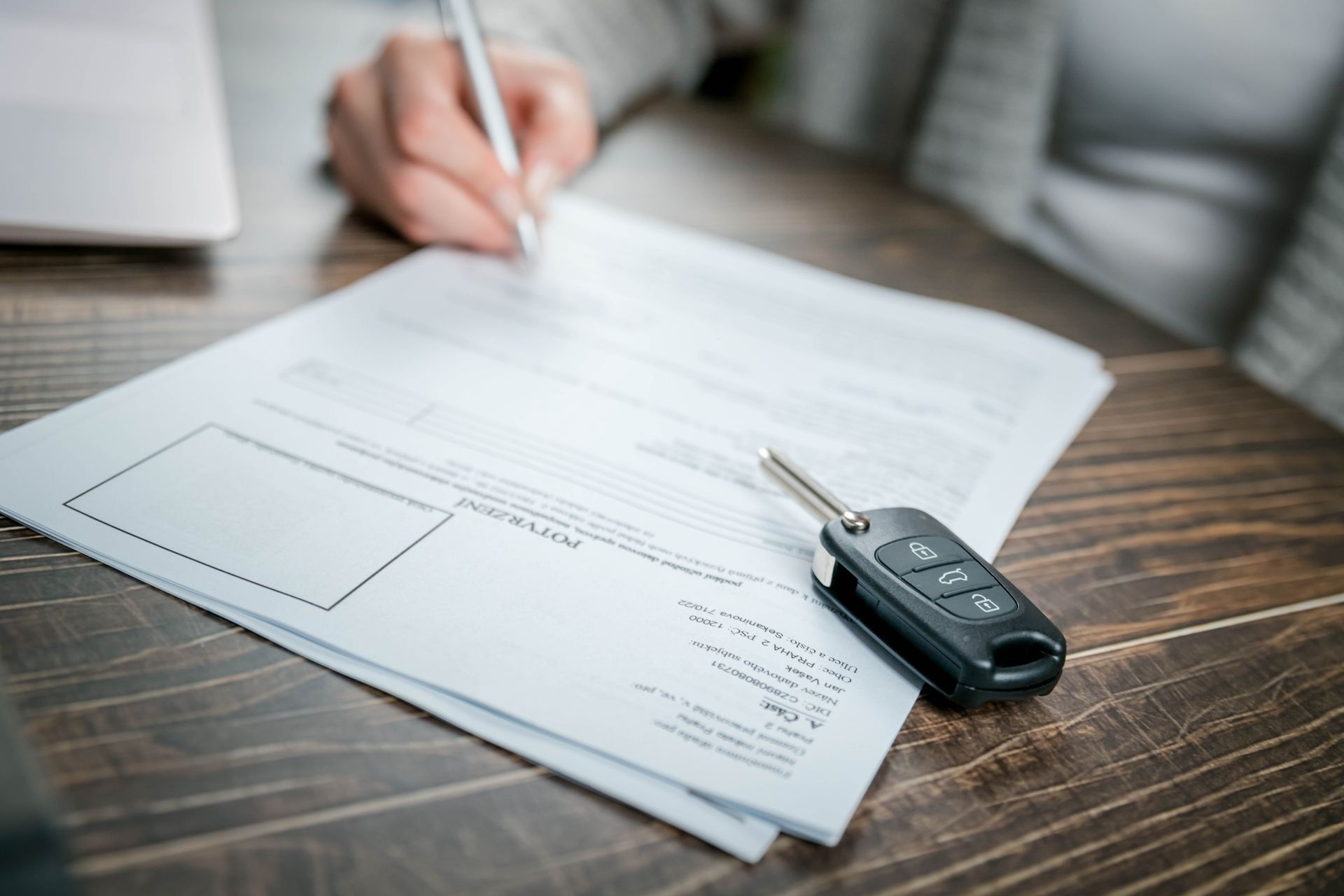 Hand signing car insurance paperwork beside car keys on a wooden table Hand signing car insurance paperwork beside car keys on a wooden table