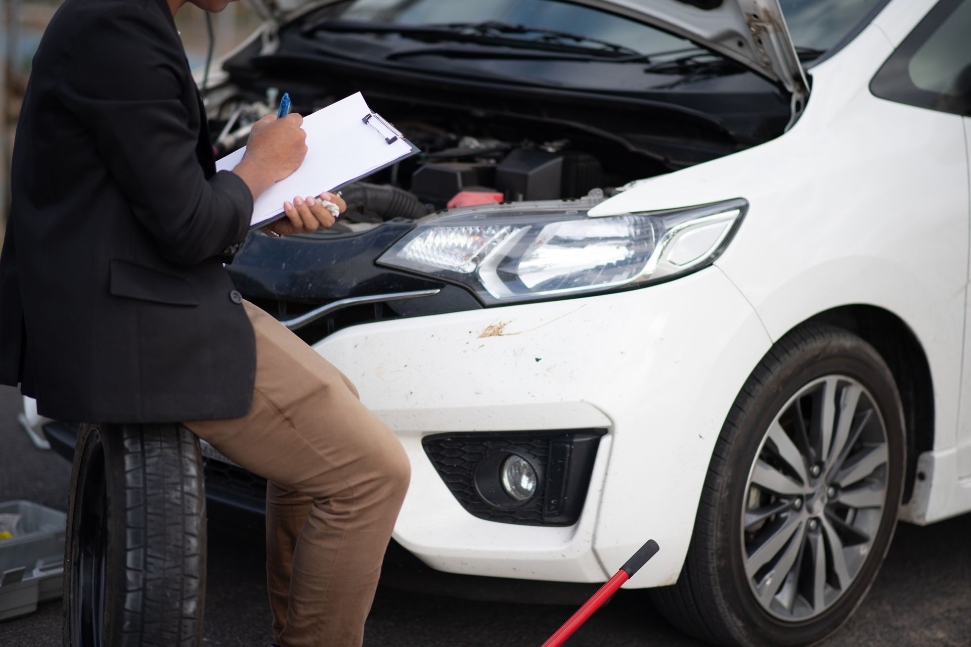 Person taking notes beside a white car with its hood open, inspecting the front end
