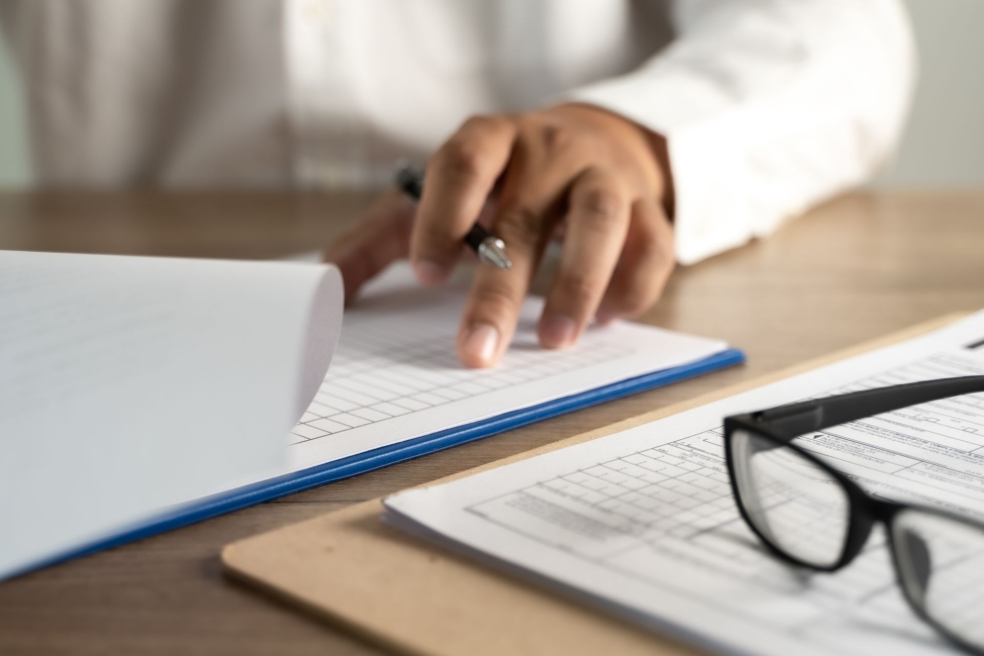 Hand pointing at a notebook beside glasses on a desk, with an open folder in the foreground