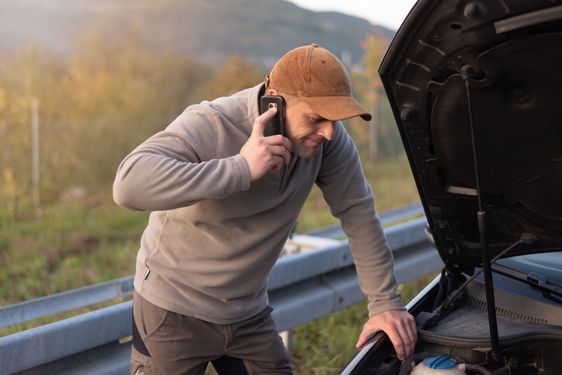 Man in an orange cap talks on a phone while inspecting an open car hood by a roadside. Man in an orange cap talks on a phone while inspecting an open car hood by a roadside.
