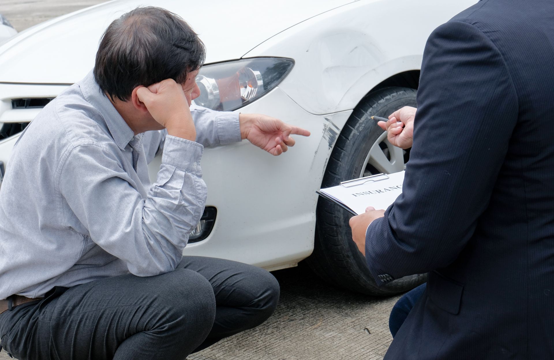 Two men inspect damage on a white car’s front wheel after a collision, one holding a clipboard.