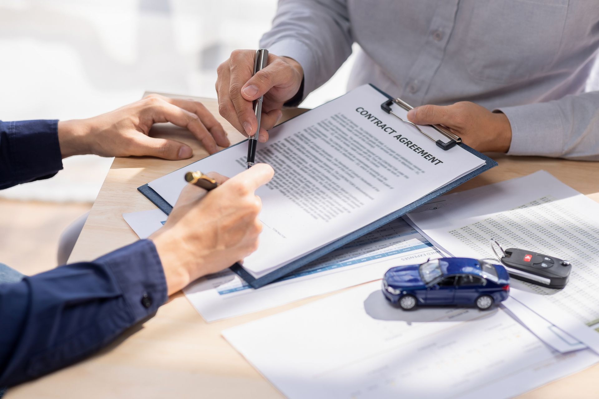 Hands reviewing and signing a car insurance document with a toy blue car and key fob on the table