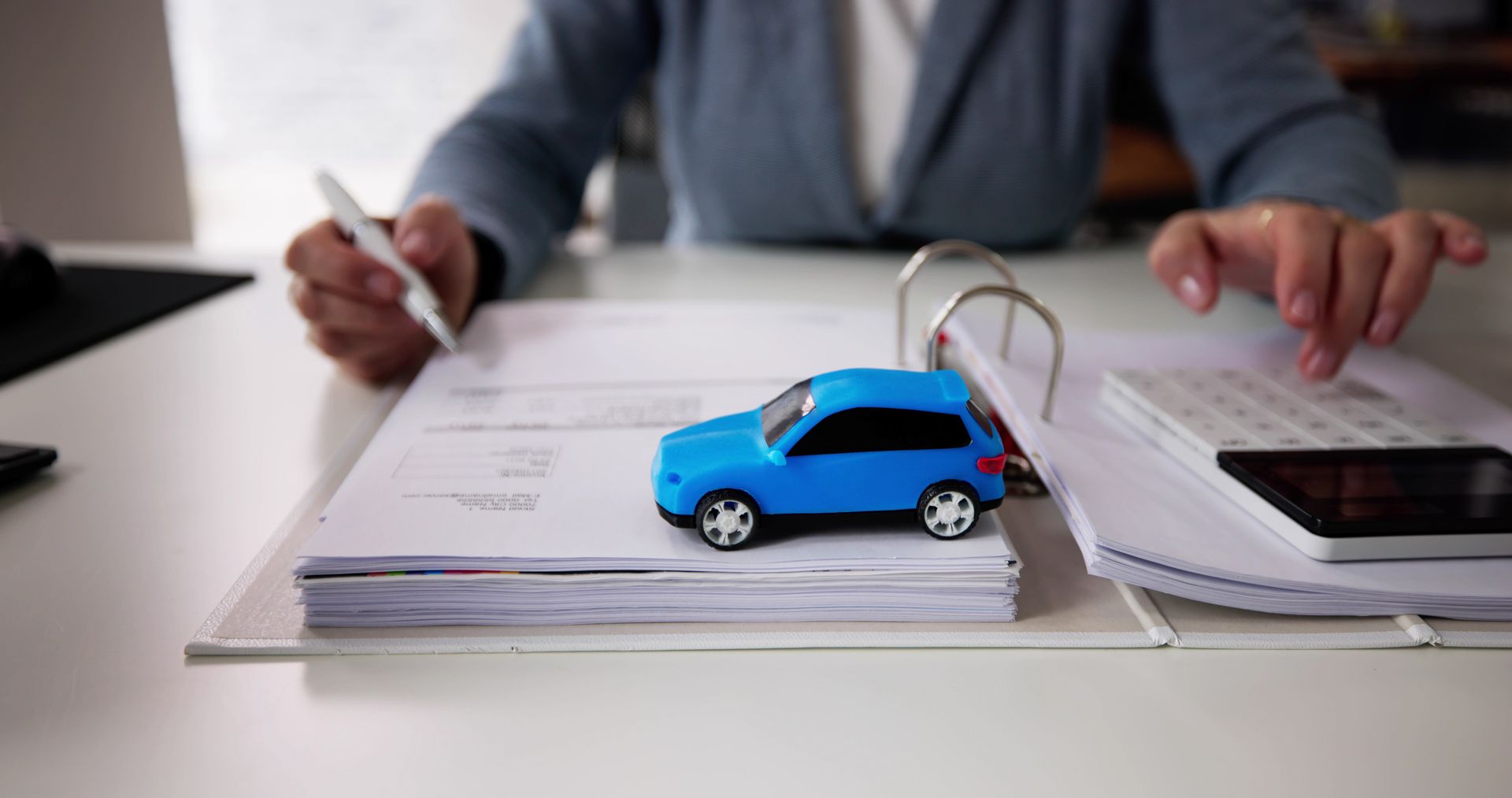 Blue toy car on stacked papers, with hands writing in the background and a smartphone nearby.