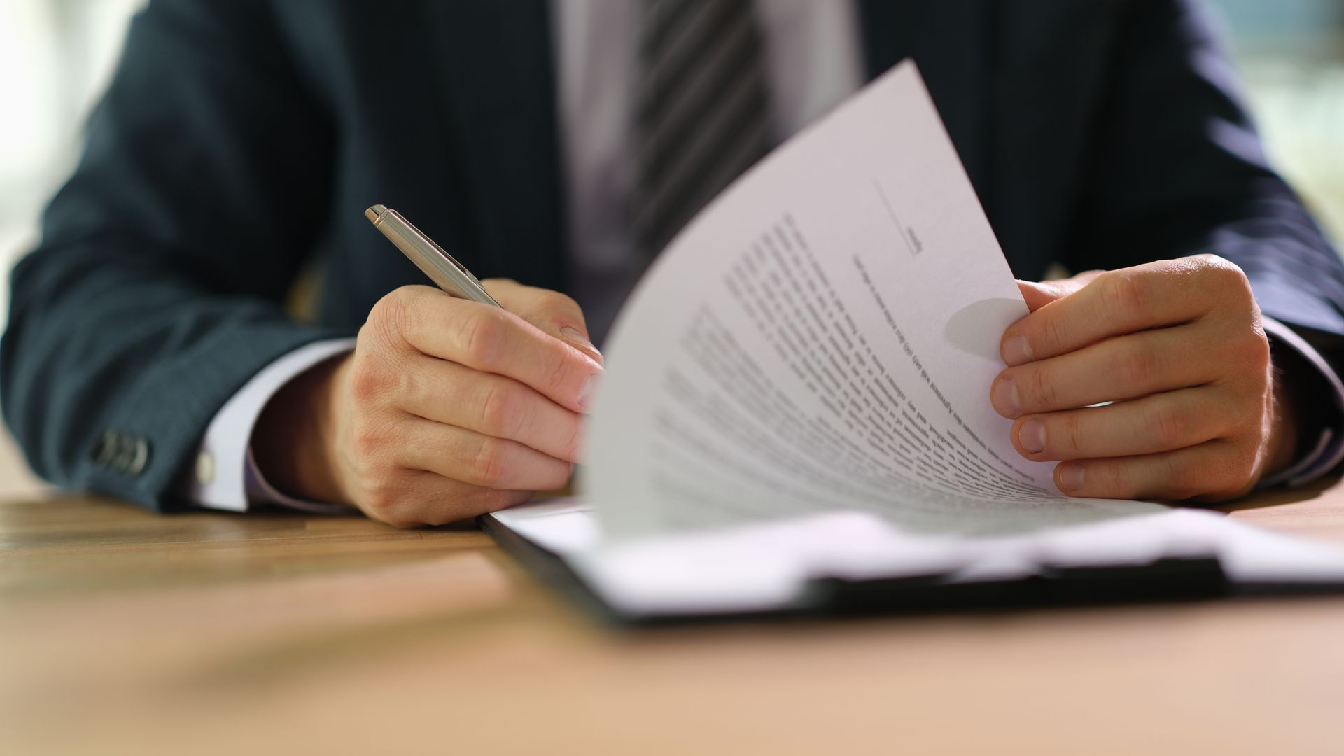 Hands holding a pen and reviewing papers at a desk, with a laptop in the foreground. Hands holding a pen and reviewing papers at a desk, with a laptop in the foreground.