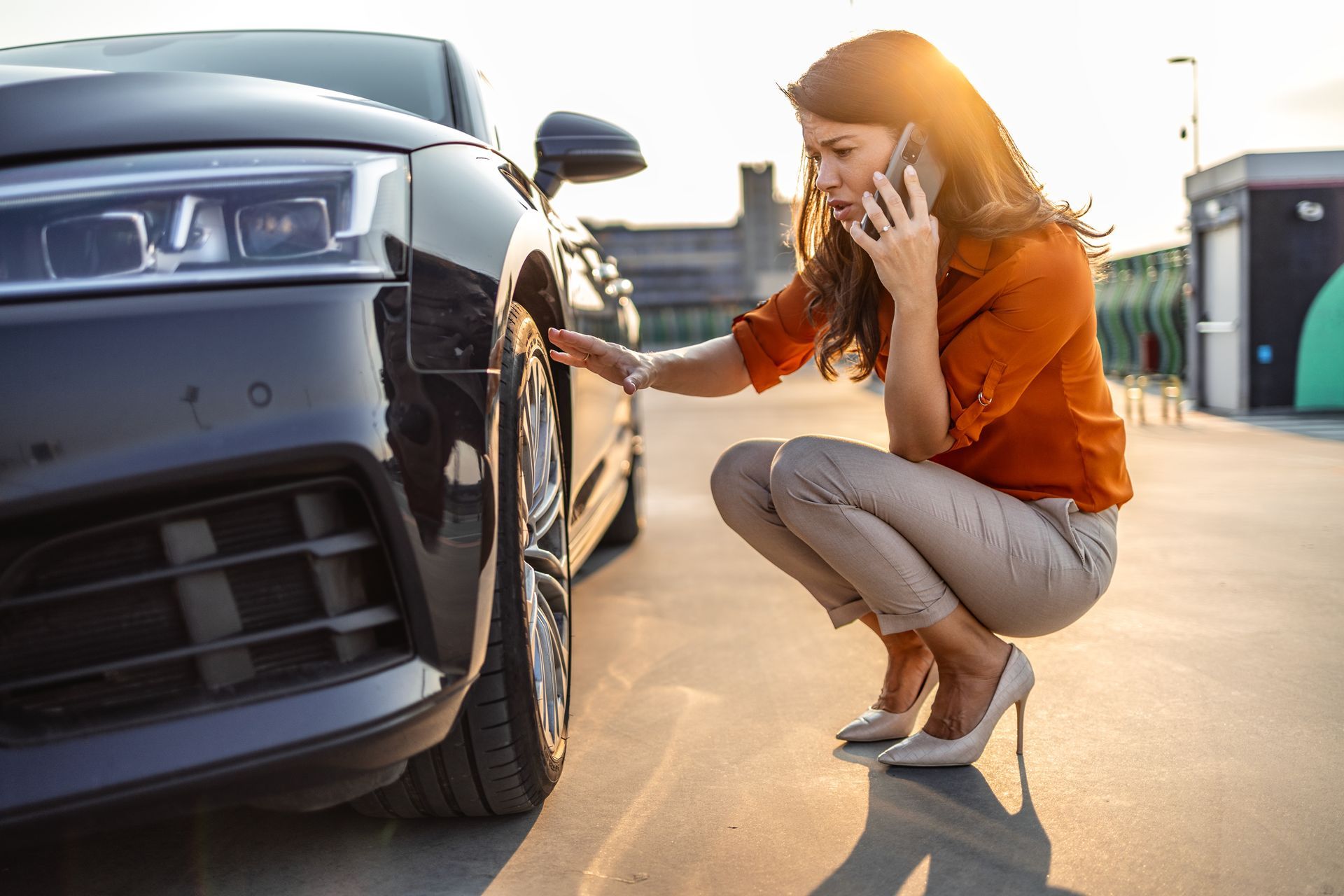 Woman crouching beside a black car, inspecting the front wheel while talking on a phone. Woman crouching beside a black car, inspecting the front wheel while talking on a phone.