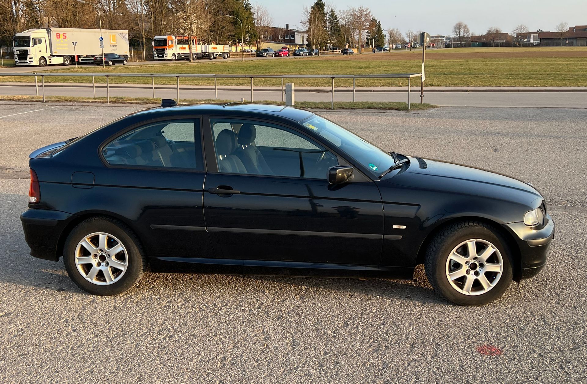Dark blue sedan parked on gravel beside a road with fields and trucks in the background Dark blue sedan parked on gravel beside a road with fields and trucks in the background