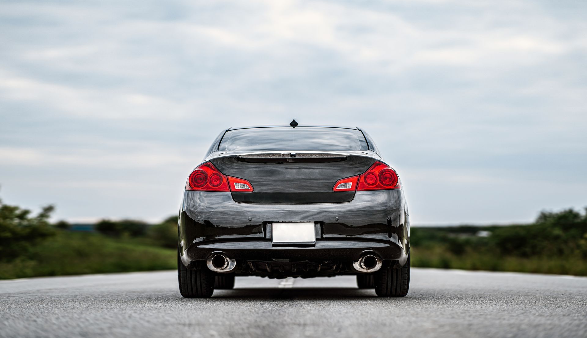 Rear view of a black sedan on a road, with red taillights and a cloudy sky overhead
