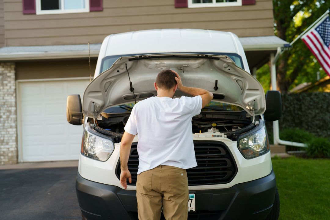 Person inspecting the open hood of a white van in a driveway with a house in the background Person inspecting the open hood of a white van in a driveway with a house in the background