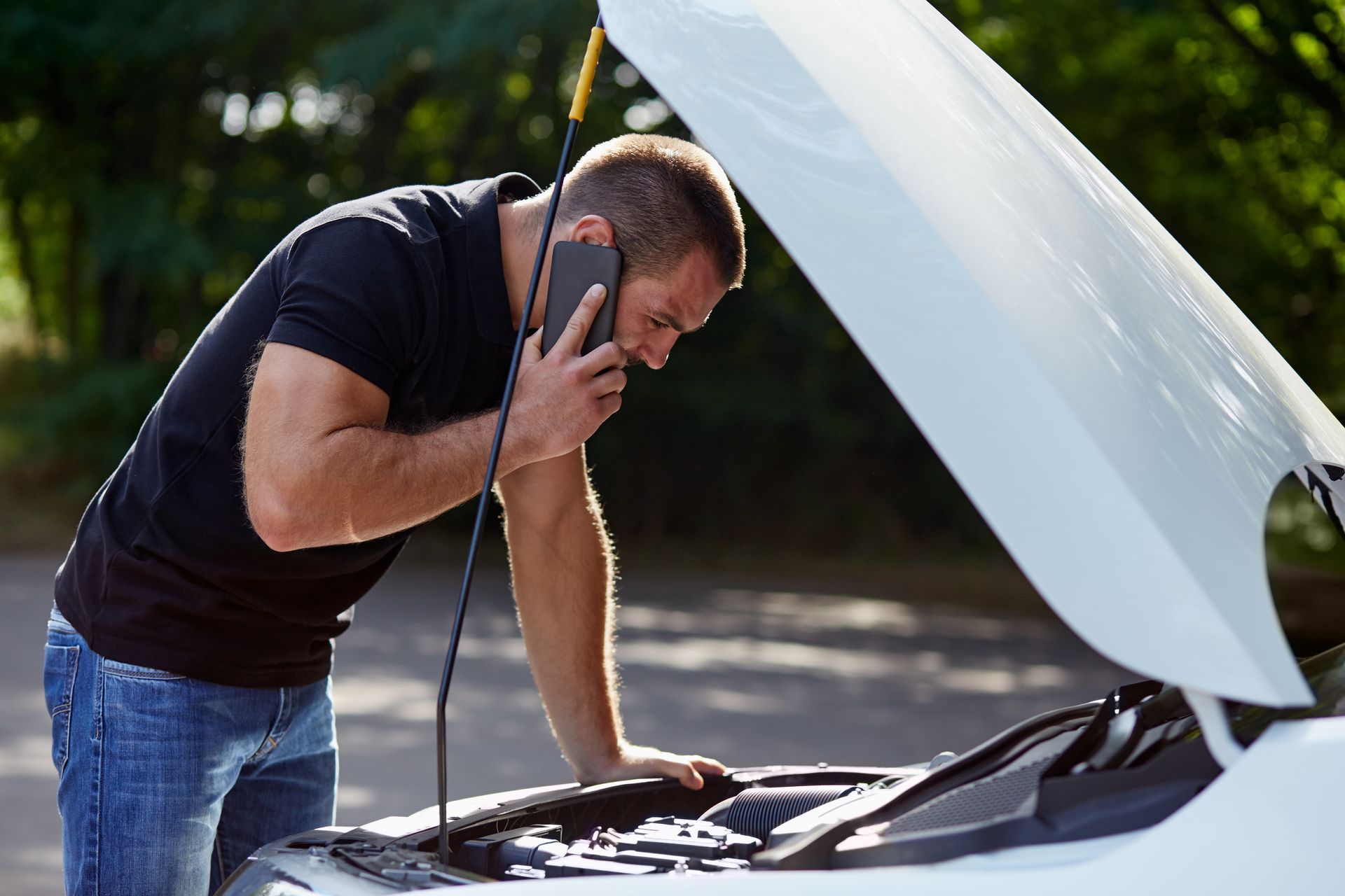 Man inspecting a car engine while holding a phone, with the hood open outdoors.