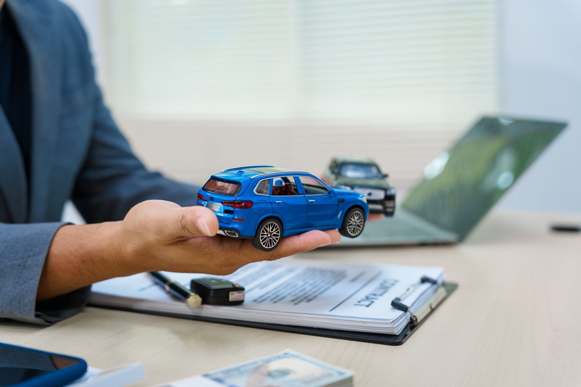Person holding a blue toy car over a clipboard with a laptop in the background