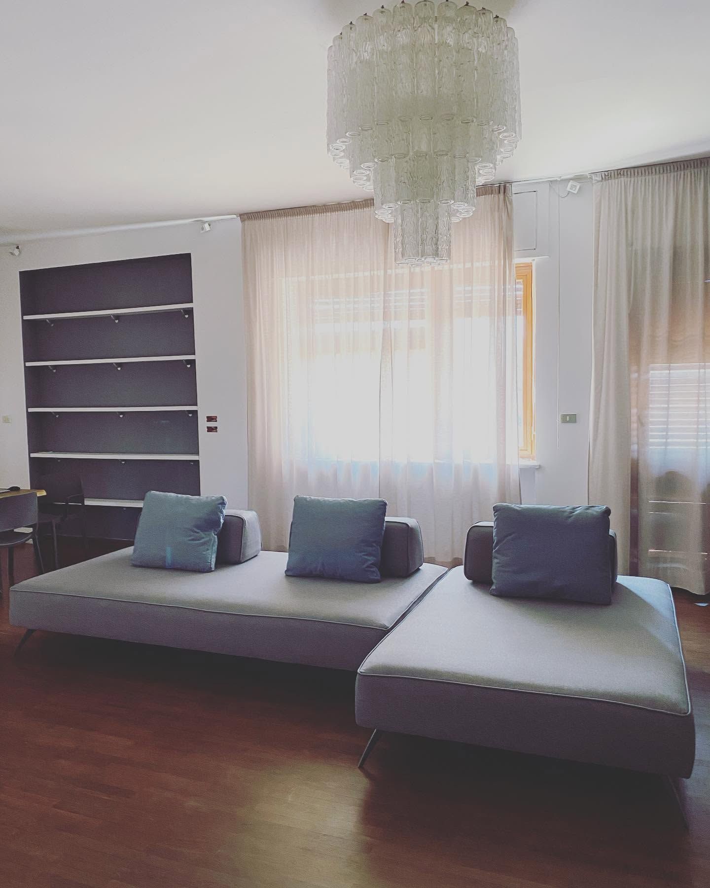 Soggiorno moderno con divano angolare grigio, cuscini blu, lampadario e grandi finestre con tende