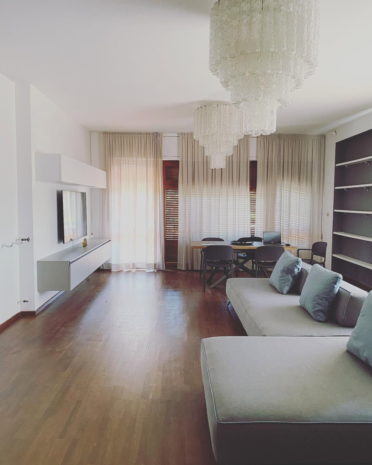 Soggiorno moderno con pavimento in legno, lampadario, divano componibile e ampie finestre con tende
