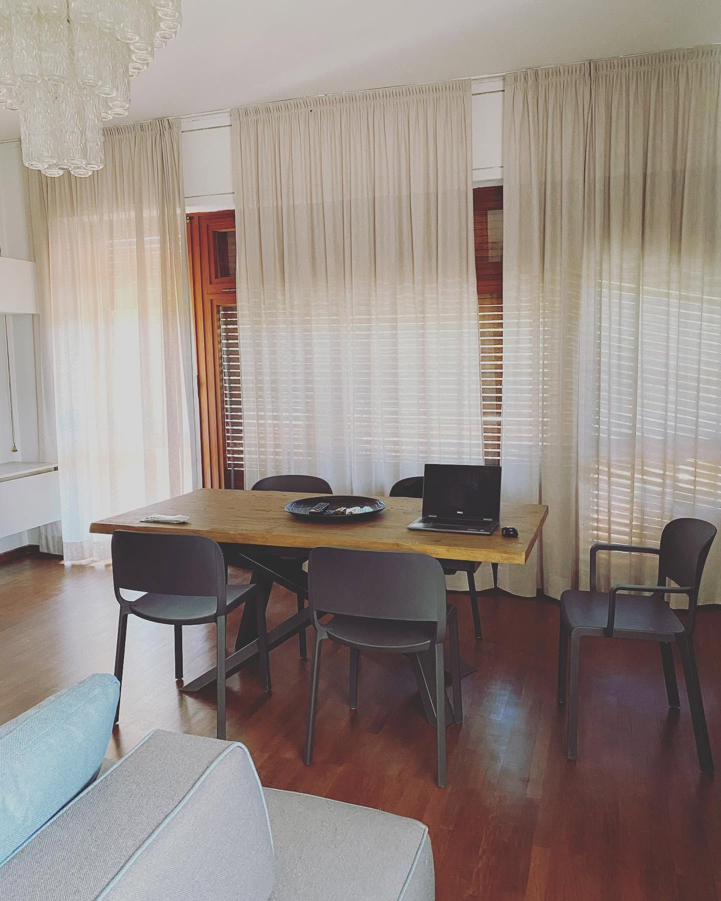 Zona pranzo con un tavolo in legno, sedie, un portatile e alte tende trasparenti accanto a luminose finestre.
