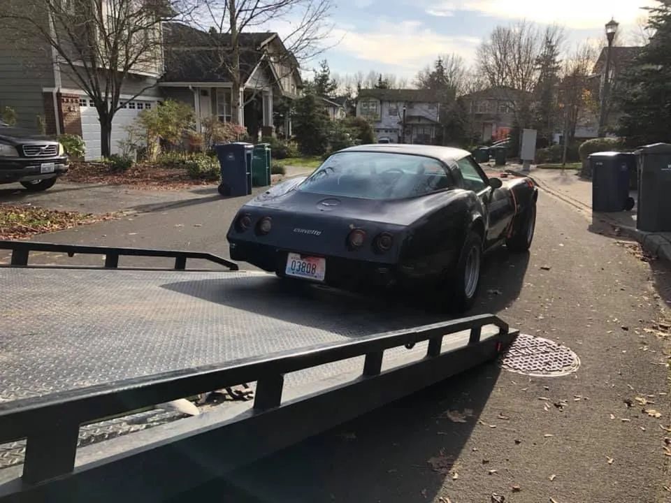 A dark-colored classic Chevrolet Corvette being loaded onto a flatbed tow truck on a suburban residential street.