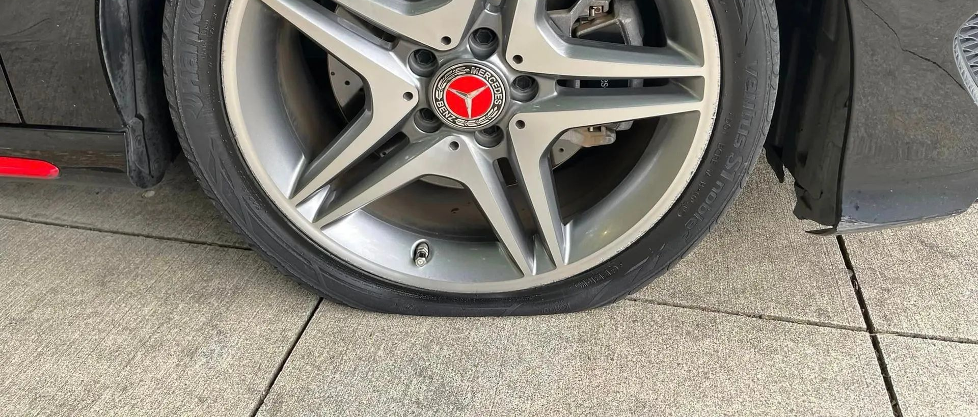 A close-up view of a silver Mercedes-Benz wheel on a car with a visibly flat tire parked on a concrete surface.