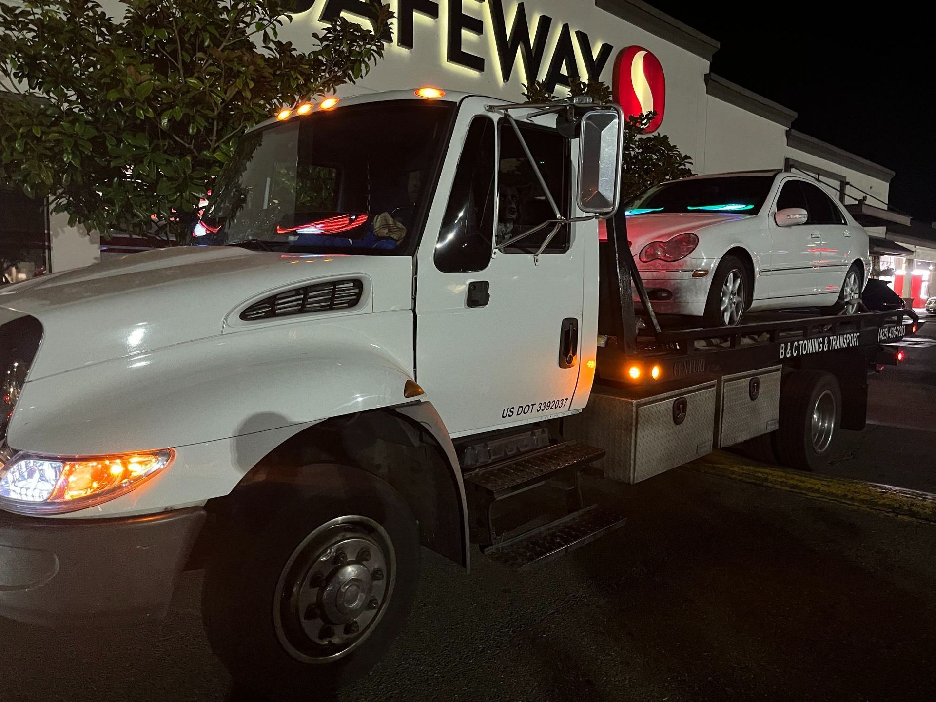 Tow truck hauling a white car at night in a Safeway parking lot