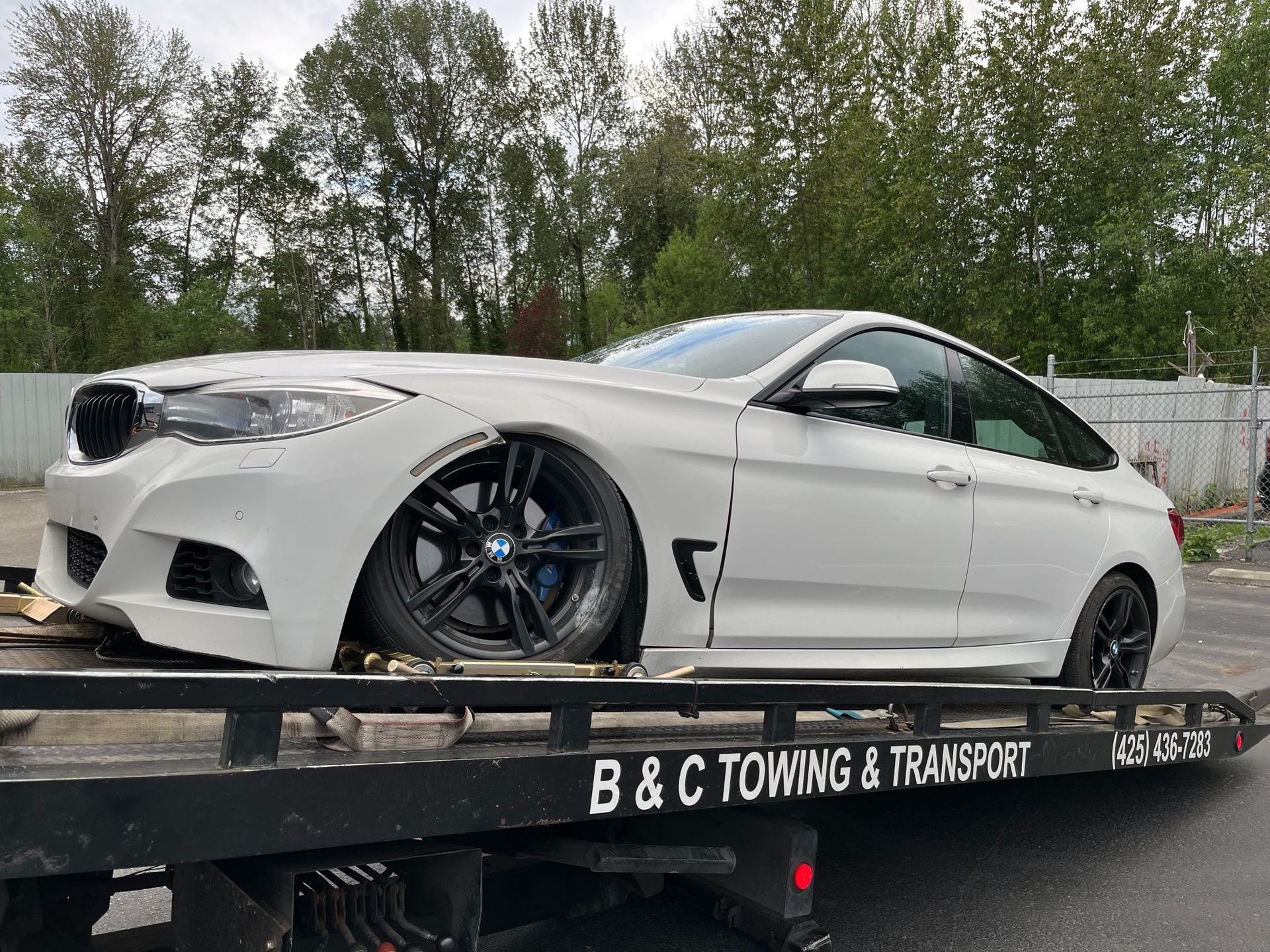 White BMW coupe on a tow truck with B&G Towing & Transport, parked outdoors near trees