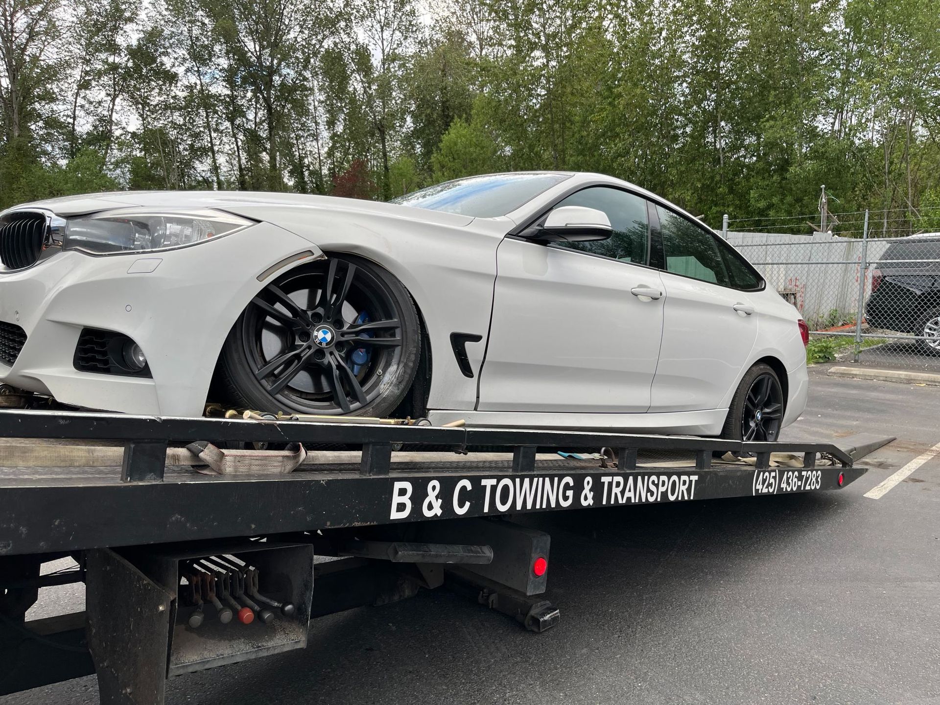 White sports coupe on a black tow truck outdoors, with trees and a fence in the background