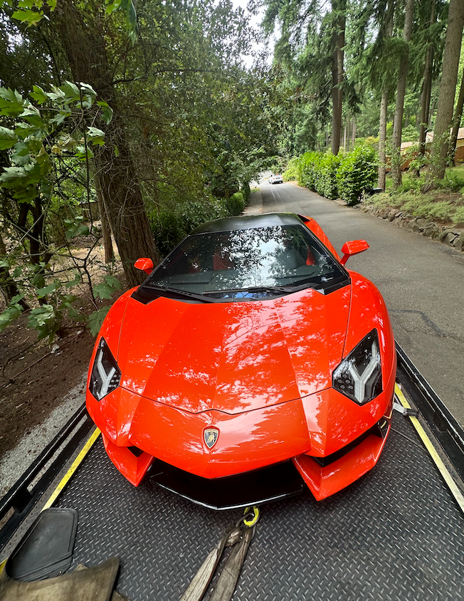 Bright orange sports car on a narrow tree-lined road, viewed from the front.