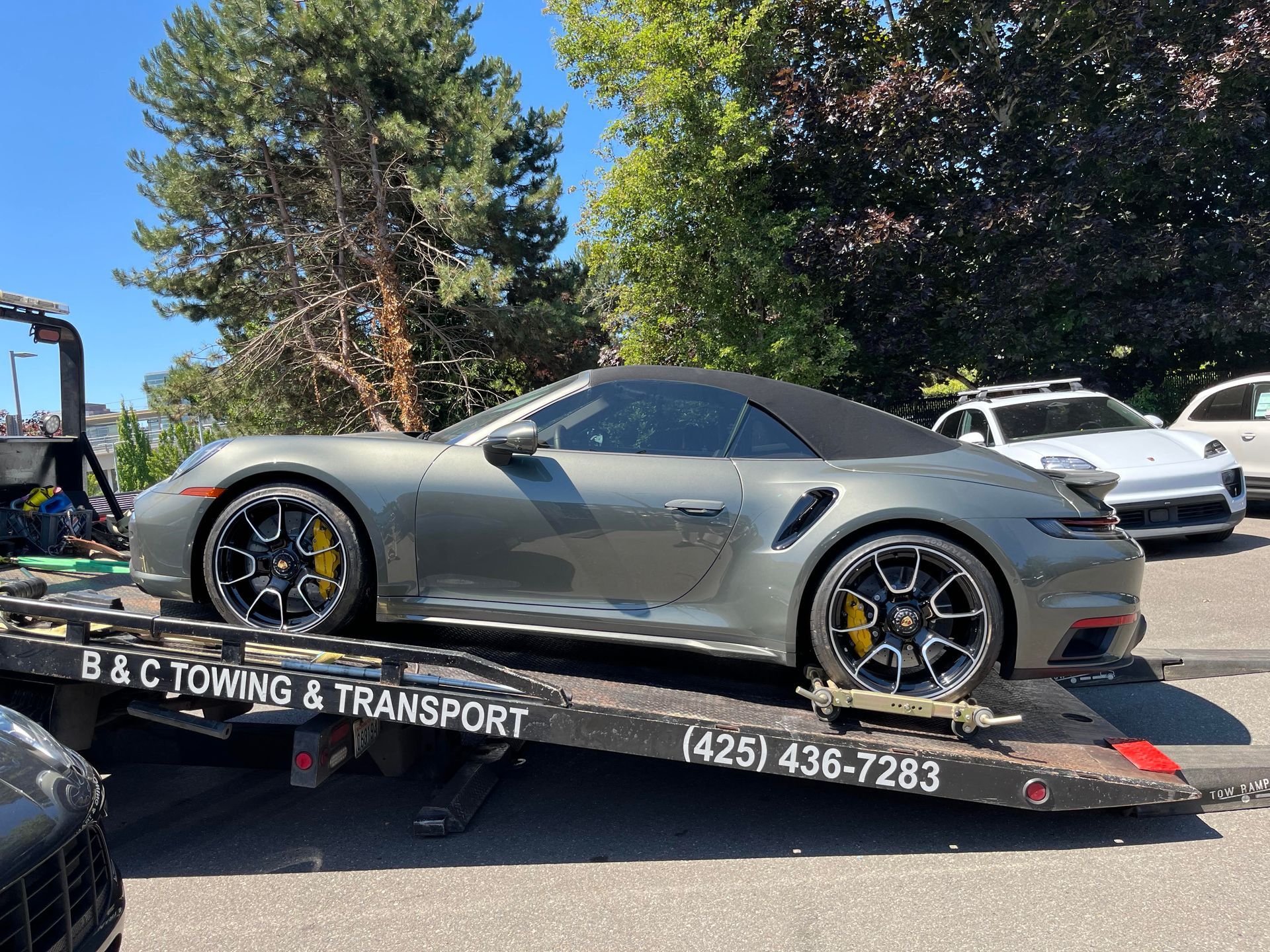 Silver sports car on a tow truck bed with a flat rear tire, parked outdoors near trees.