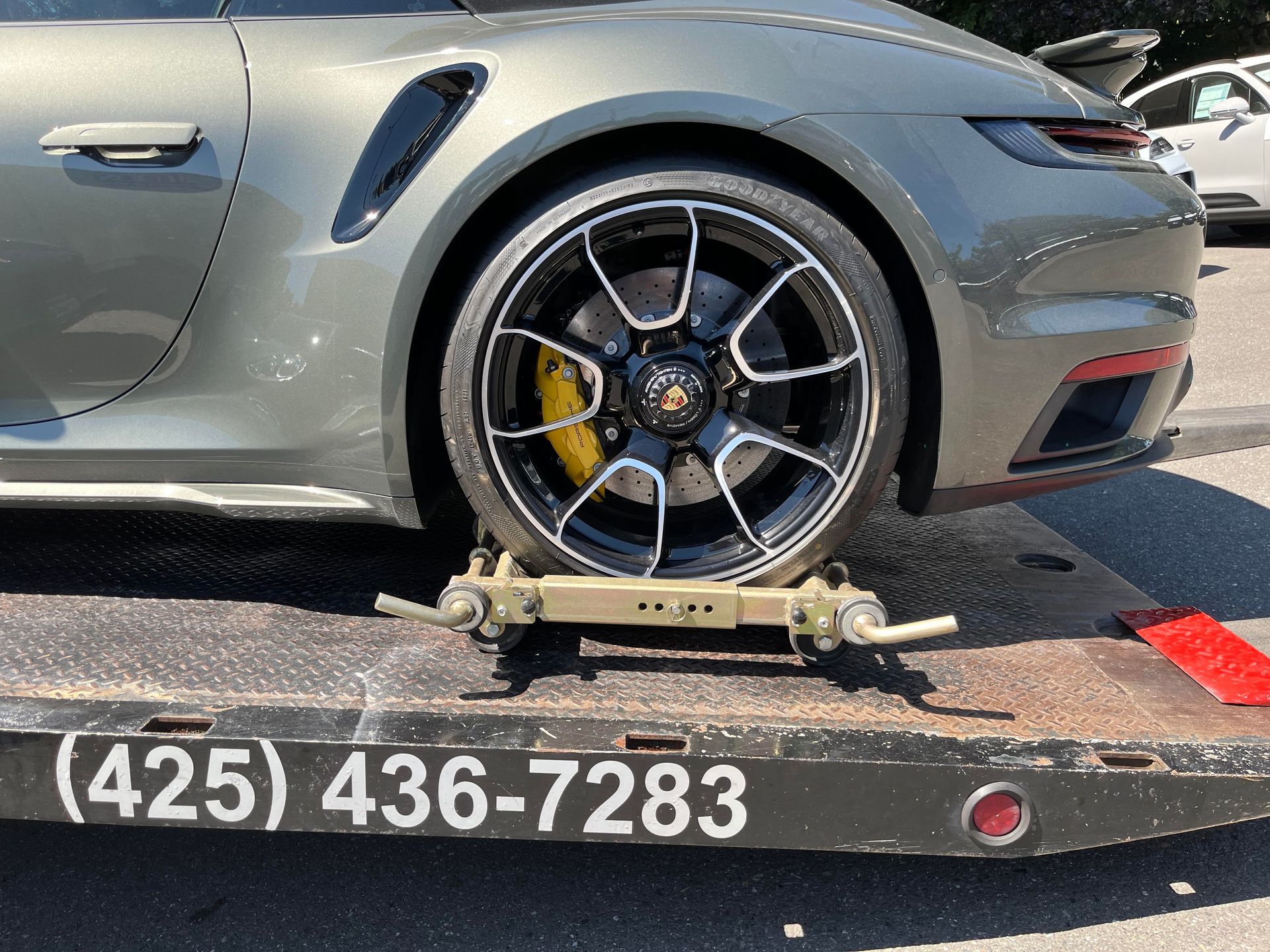 Gray sports car with front wheel on a tow dolly, roadside scene with phone number on the bumper.