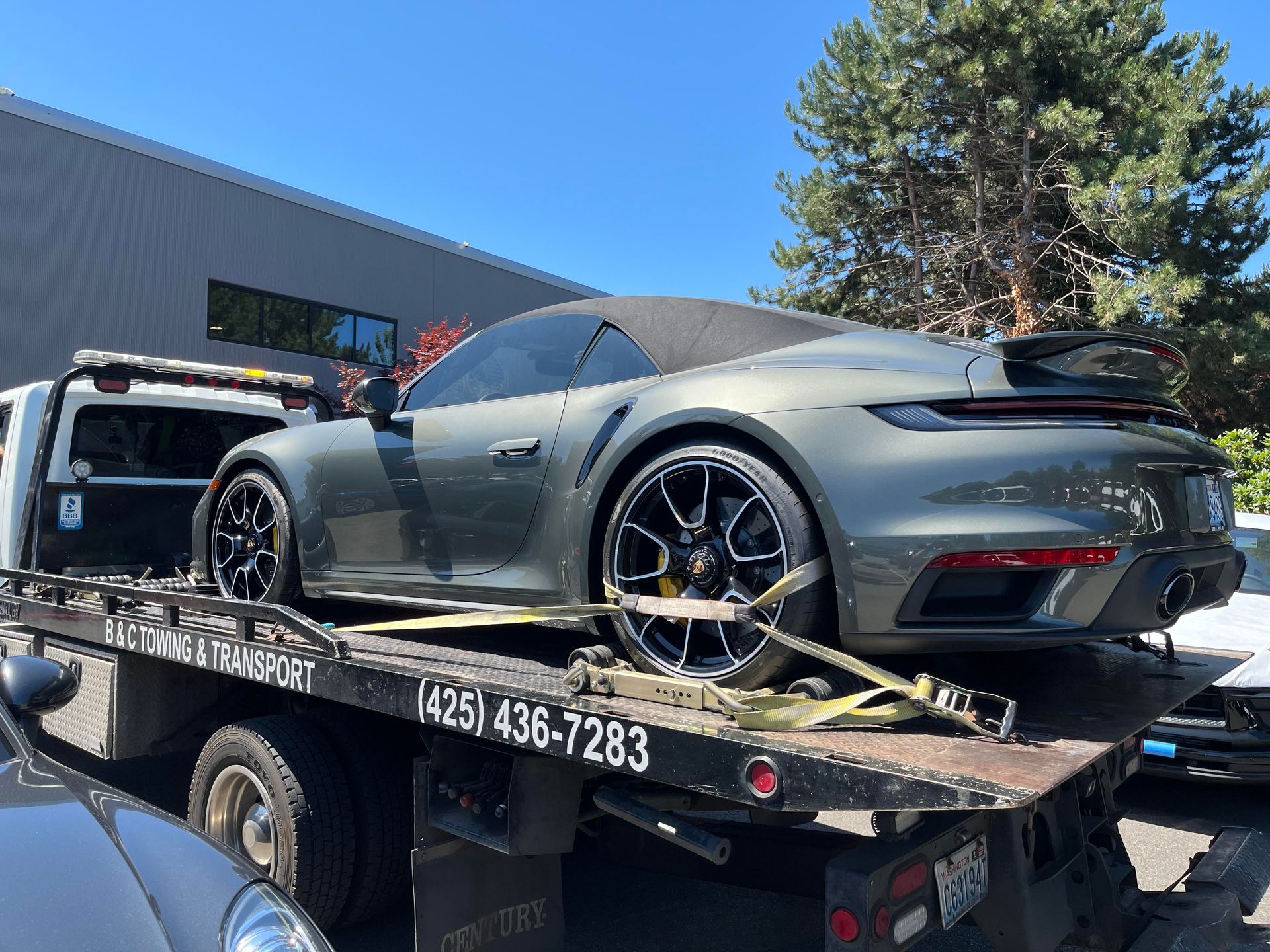 Gray sports car strapped on a tow truck in front of a building under a clear blue sky.