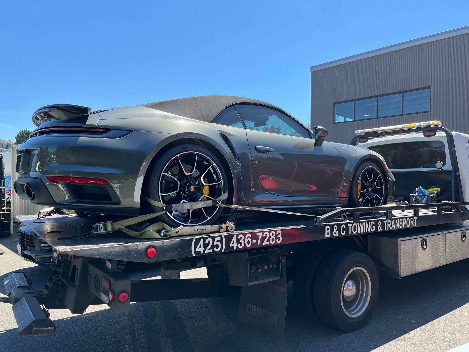 Gray sports car on a tow truck bed under a clear blue sky, with a flatbed and tow equipment visible.