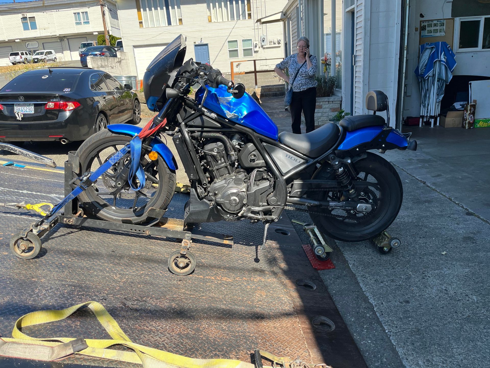 Blue motorcycle on a dolly beside a garage, with a person standing nearby and a car in the background