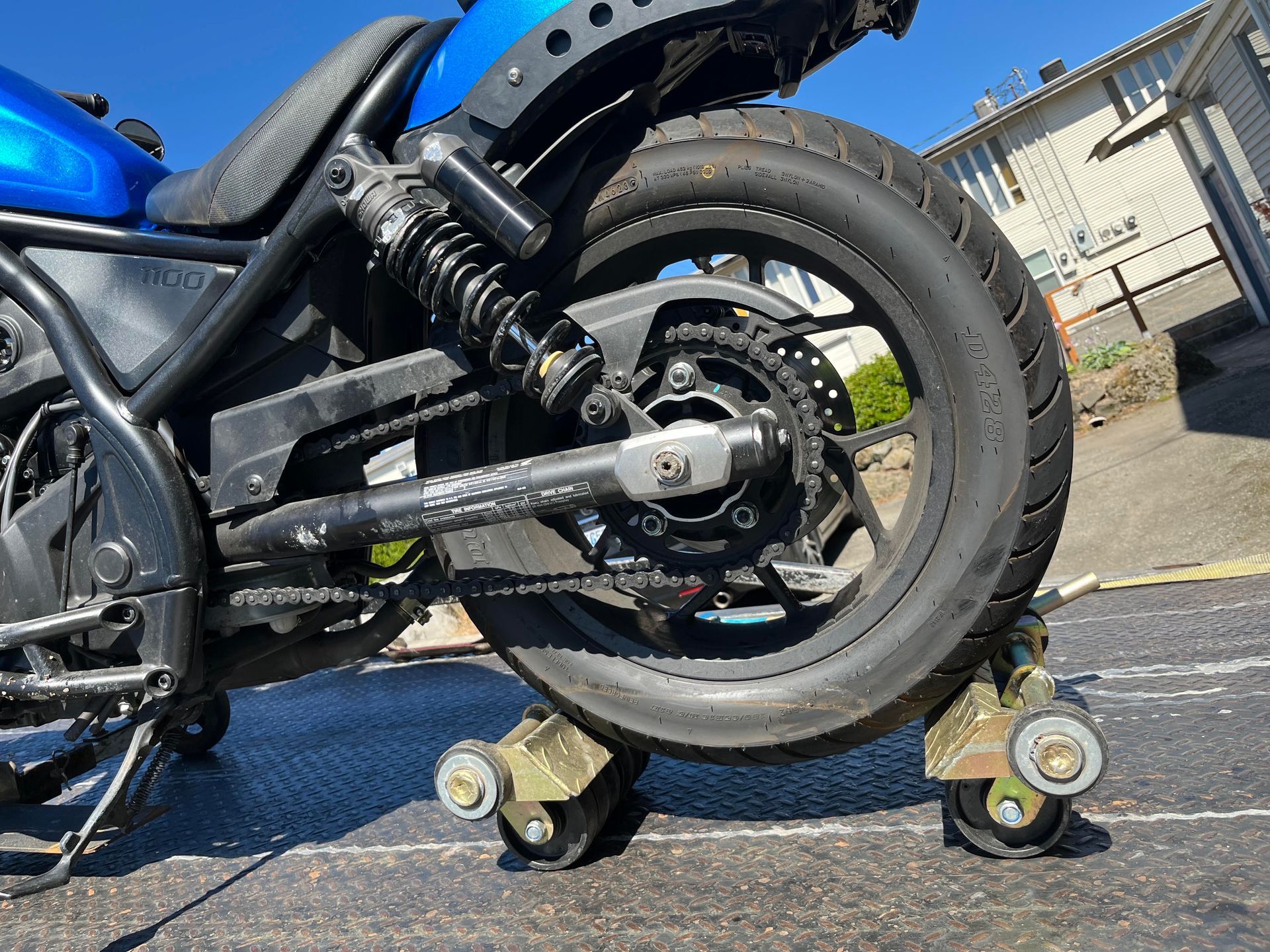 Motorcycle rear wheel lifted on small training stands outdoors, with a blue bike body and driveway background.