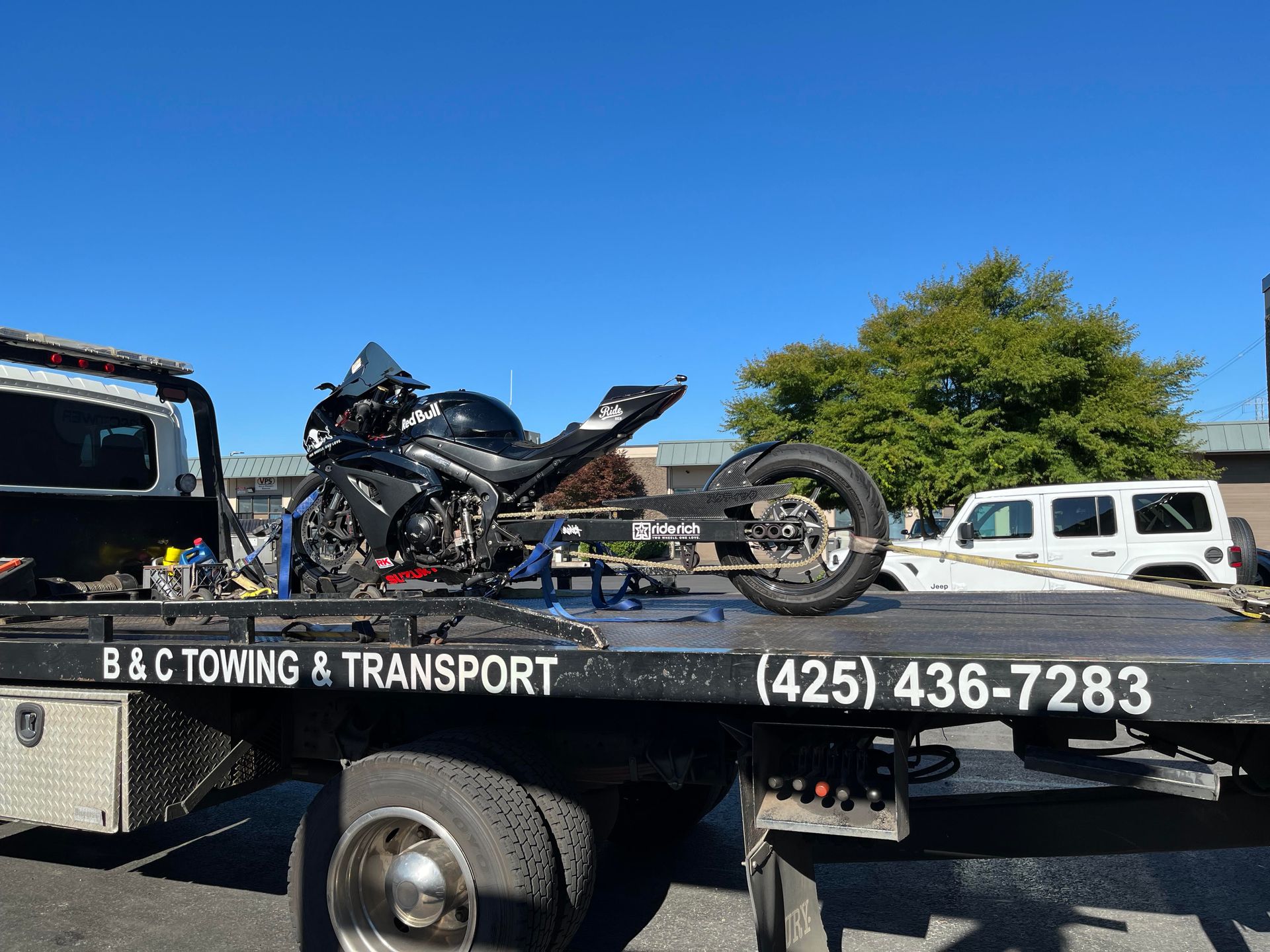 Motorcycle on a tow truck flatbed under a clear blue sky, with “B&C Towing & Transport” visible on the side.