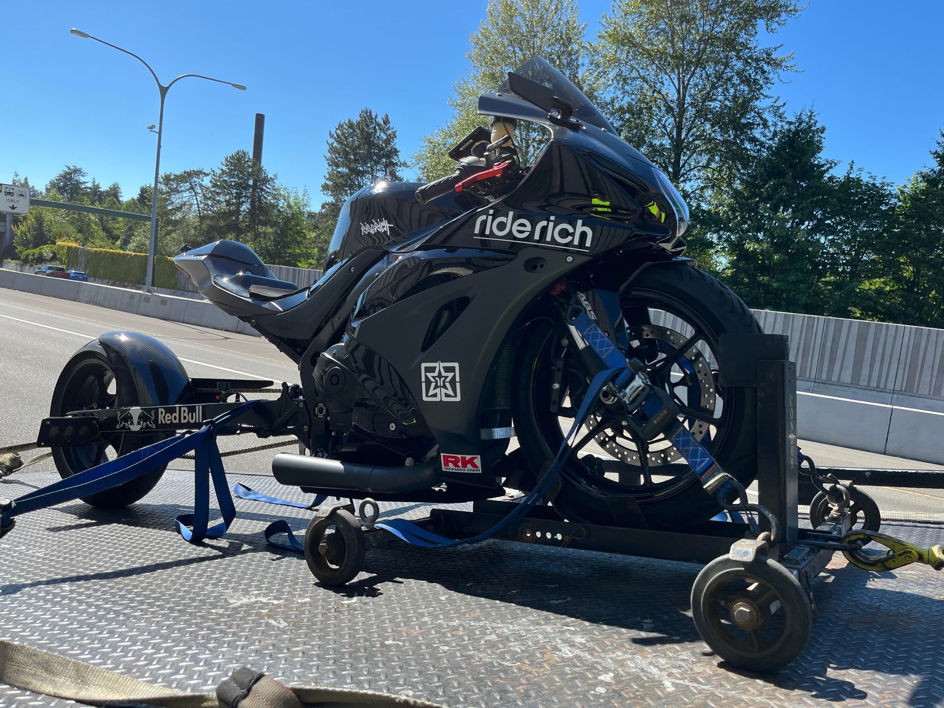 Black sport motorcycle strapped on a trailer in bright daylight, parked on a road near trees.