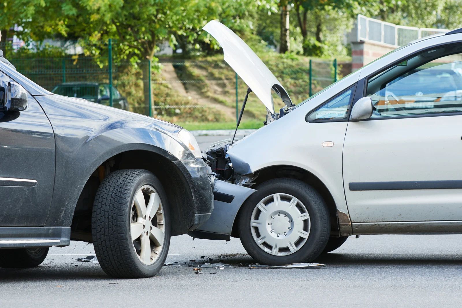 Two cars collide on a street, with the white car’s hood raised after impact.