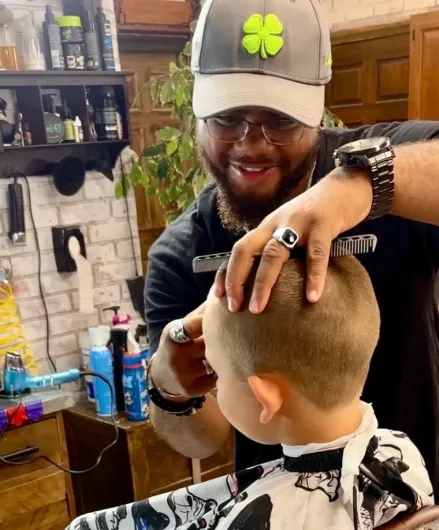 A stylist with brown hair cuts the long hair of a client wearing a black cape in a barbershop with a brick wall.