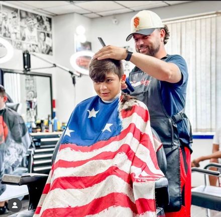 A stylist with brown hair cuts the long hair of a client wearing a black cape in a barbershop with a brick wall.