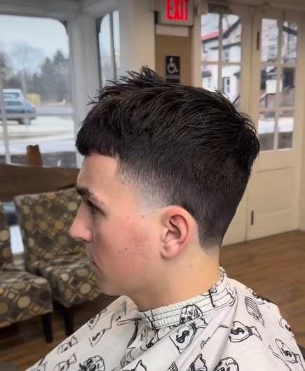 A person with a dark, textured short haircut and a faded side profile, seated in a barber chair.