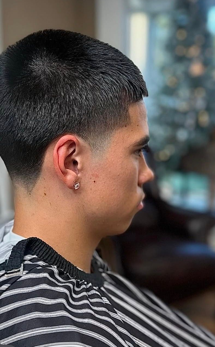 A stylist with brown hair cuts the long hair of a client wearing a black cape in a barbershop with a brick wall.